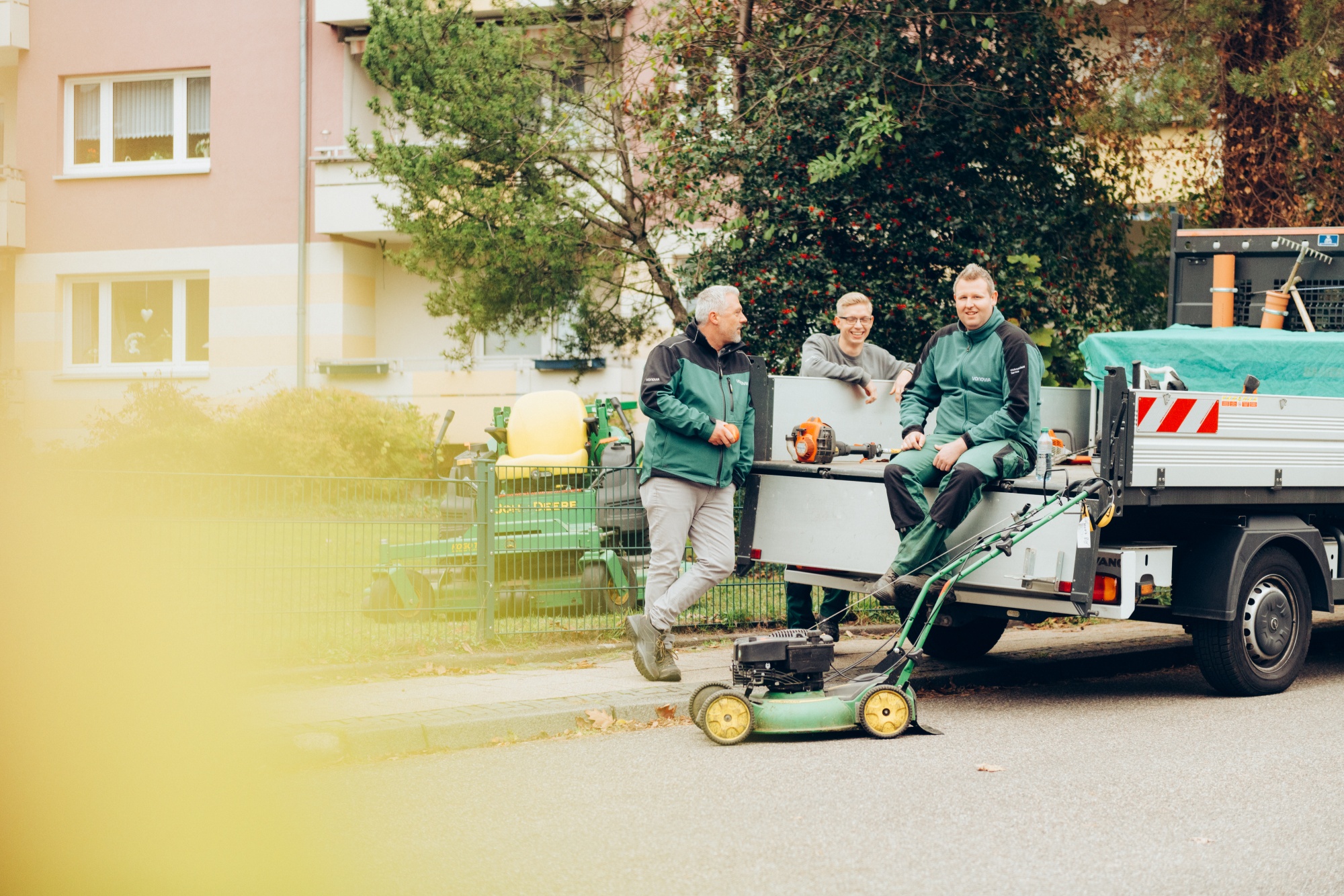 Vonovia Mitarbeitende des Grünflächen- und Gebäudeservices lachend auf einem Transporter mit Werkzeugen sitzend. Vonovia Mitarbeitende des Grünflächen- und Gebäudeservices lachend auf einem Transporter mit Werkzeugen sitzend.