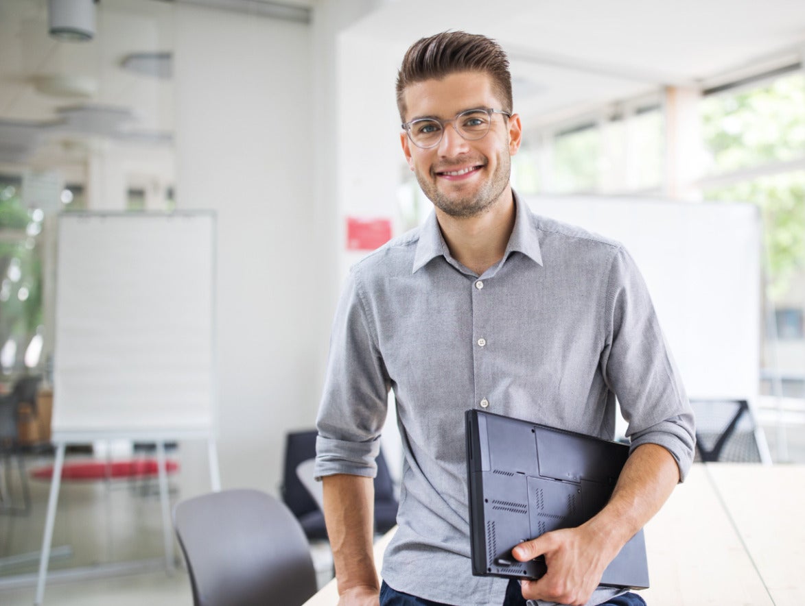 Ein junger, lächelnder Mann mit Brille und grauem Hemd steht in einem hellen, modernen Büro oder Konferenzraum. Er lehnt an einem großen Holztisch und hält einen geschlossenen, schwarzen Laptop unter dem Arm. Im Hintergrund sind große Fenster, Stühle und ein Whiteboard zu sehen.