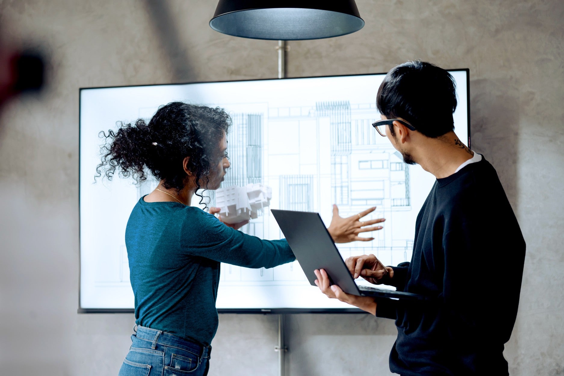 Zwei Personen besprechen ein Architekturprojekt. Eine Frau mit lockigem Haar hält ein weißes Modellbau-Element und zeigt auf eine technische Bauzeichnung, die auf einem großen Bildschirm im Hintergrund zu sehen ist. Ein Mann mit Brille und schwarzem Pullover schaut ebenfalls zum Bildschirm und hält einen geöffneten Laptop in den Händen.