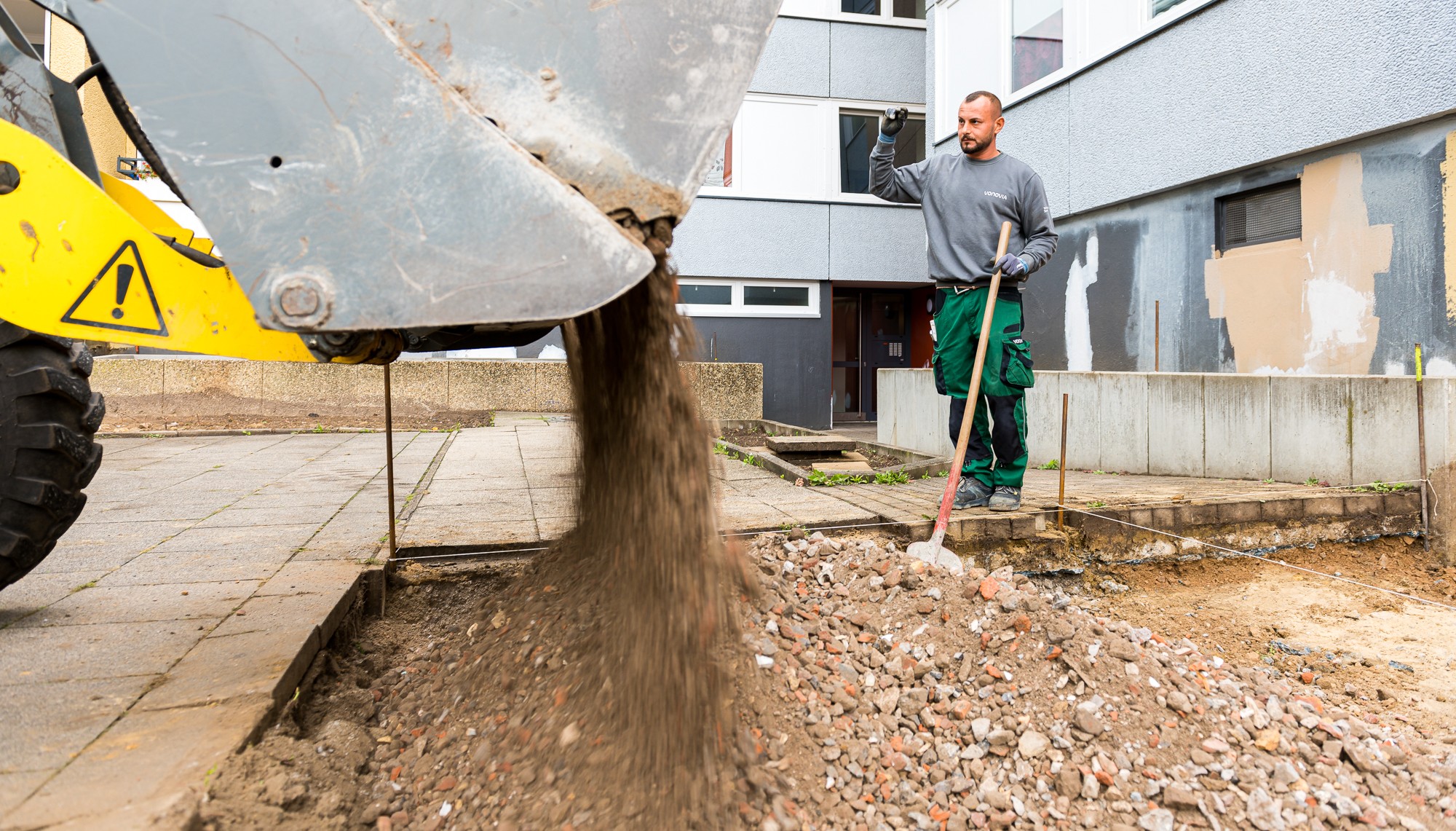 Vonovia Mitarbeiter vor einem Frontlader gibt ein Zeichen an den Fahrer zur Auffüllung eines Fundaments. Vonovia Mitarbeiter vor einem Frontlader gibt ein Zeichen an den Fahrer zur Auffüllung eines Fundaments.