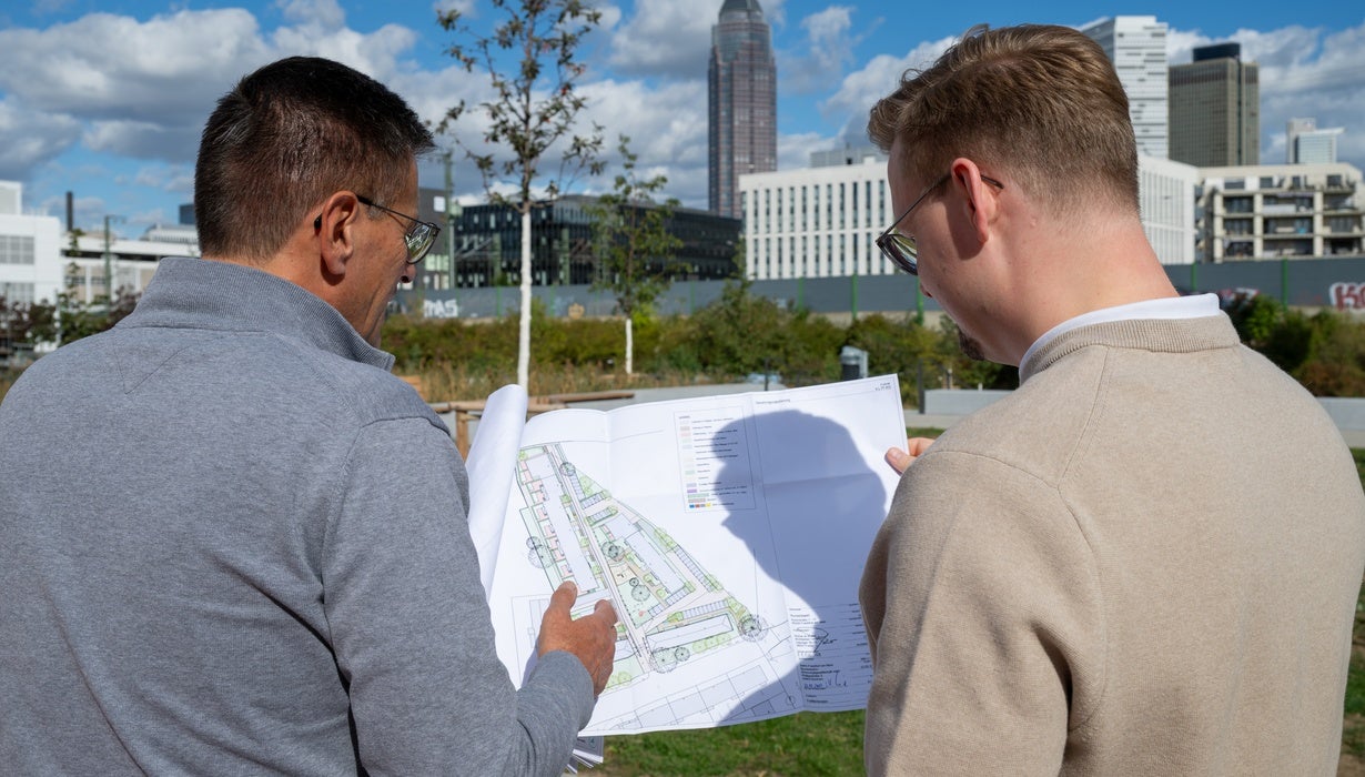 Zwei Männer blicken im Freien gemeinsam auf einen ausgebreiteten Lageplan vor einer städtischen Skyline mit Hochhäusern.